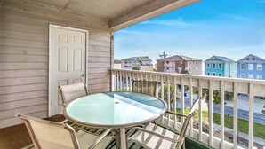 A balcony with a glass table and chairs, a white door, and a view of colorful houses. - Surfside Beach steps away from beach 8/11-8/15; 8/22-8/28; 9/8-9/20; 10/4-10/30 (Surfside Beach)
