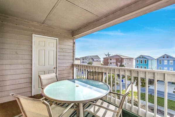 A balcony with a glass table and chairs, a white door, and a view of colorful houses. - Surfside Beach steps away from beach 8/11-8/15; 8/22-8/28; 9/8-9/20; 10/4-10/30 (Surfside Beach)