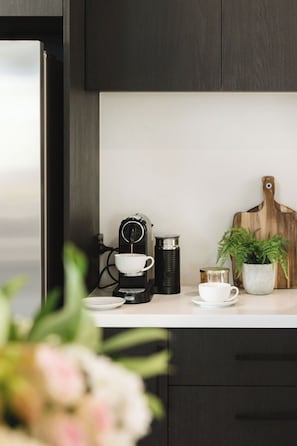 Una encimera de cocina con una cafetera, una taza, un termo, una tabla de cortar y una planta en maceta.