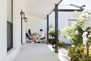 Een overdekt terras met zitplaatsen voor twee personen, omringd door witte bloemen en groen.