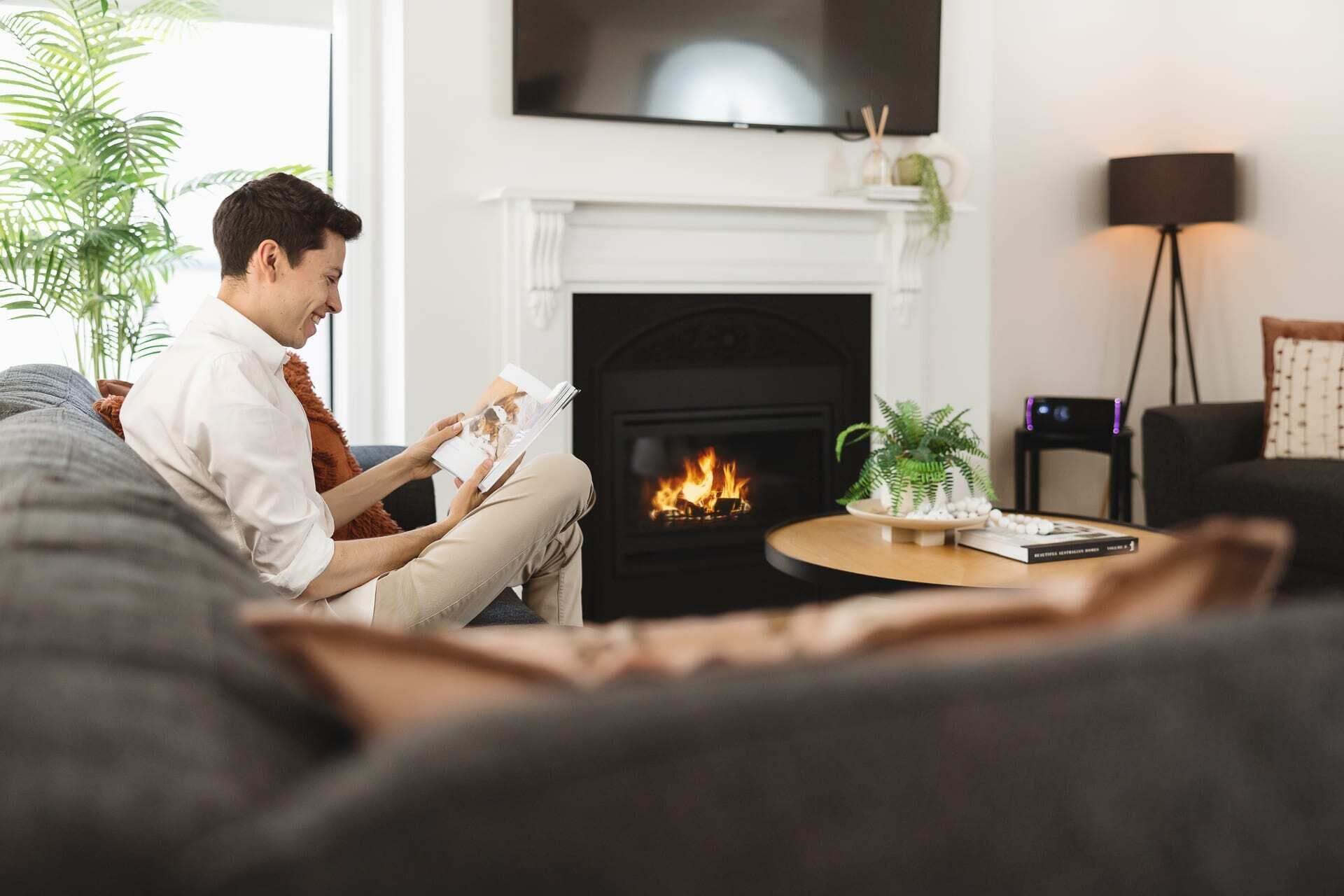 A person sitting on a couch reading a book in a living room with a fireplace, a TV mounted on the wall, and a floor lamp.