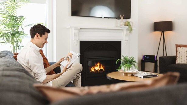Una persona sentada en un sofá leyendo un libro en una habitación con chimenea, un televisor montado encima y una pequeña mesa con una planta y un libro.