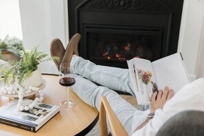 Una persona descansando en un sofá con un libro y una copa de vino tinto sobre una mesa, con una chimenea al fondo.