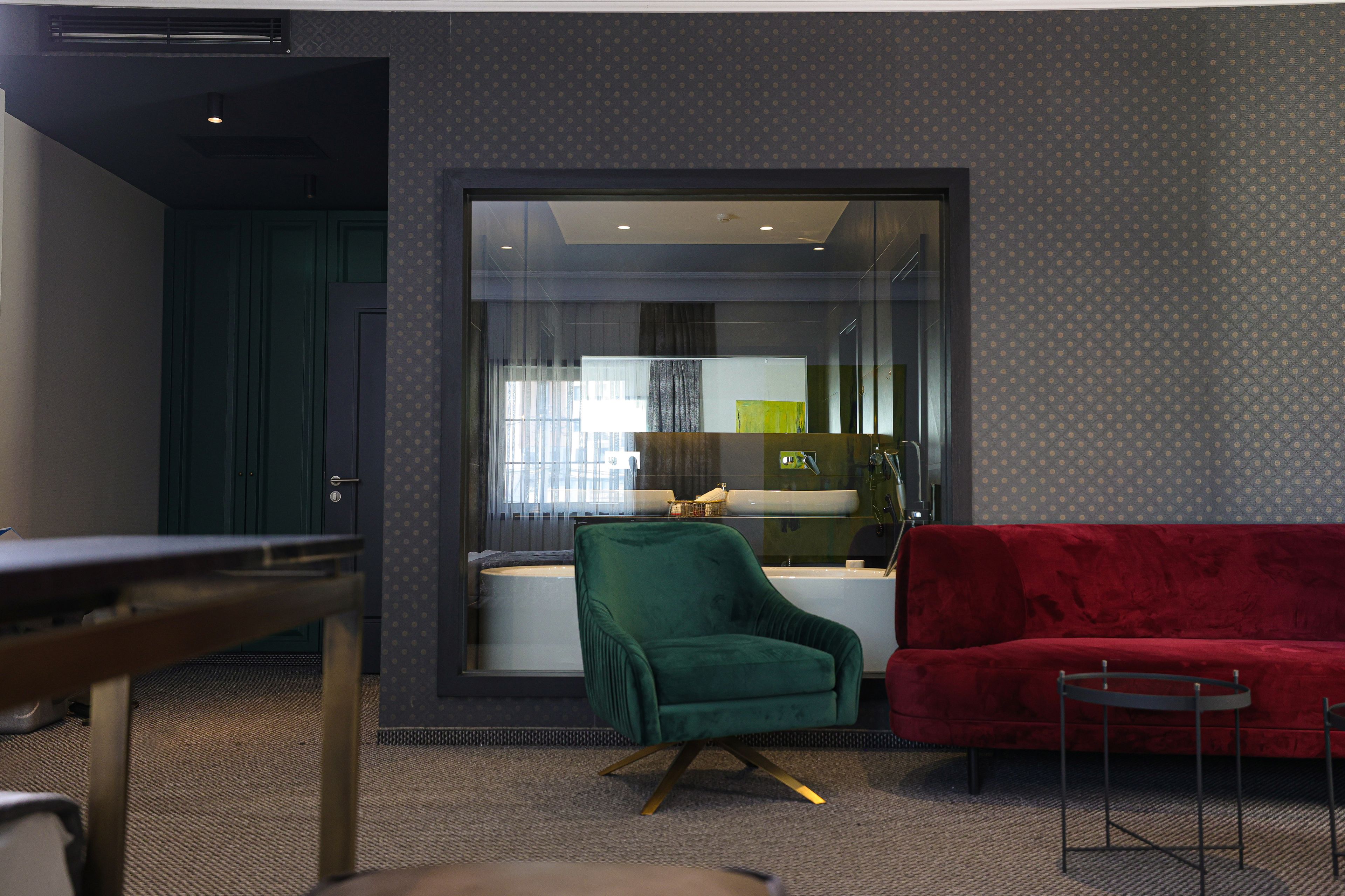 Una sala de estar moderna con un sofá rojo, un sillón verde y una pequeña mesa. Un separador de vidrio divide la zona de estar de una cocina.