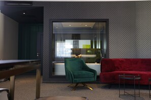 Una sala de estar moderna con un sofá rojo, un sillón verde y una pequeña mesa. Un separador de vidrio divide la zona de estar de una cocina.