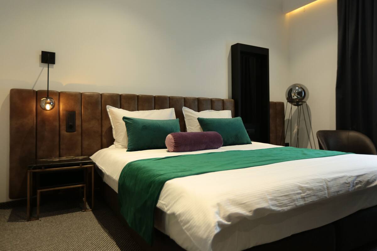 A hotel room with a large bed, a nightstand, a wall-mounted lamp, and a chair.