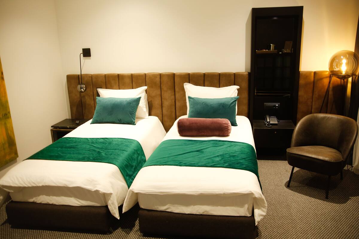 A hotel room with two beds, a brown upholstered headboard, a nightstand with a lamp, and a chair.