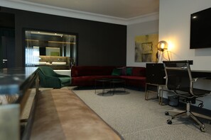 A modern hotel room with a red sofa, green armchair, and a small round table. There is a TV mounted on the wall and a lamp on the floor. - Hotel Rubis (Ferizaj)