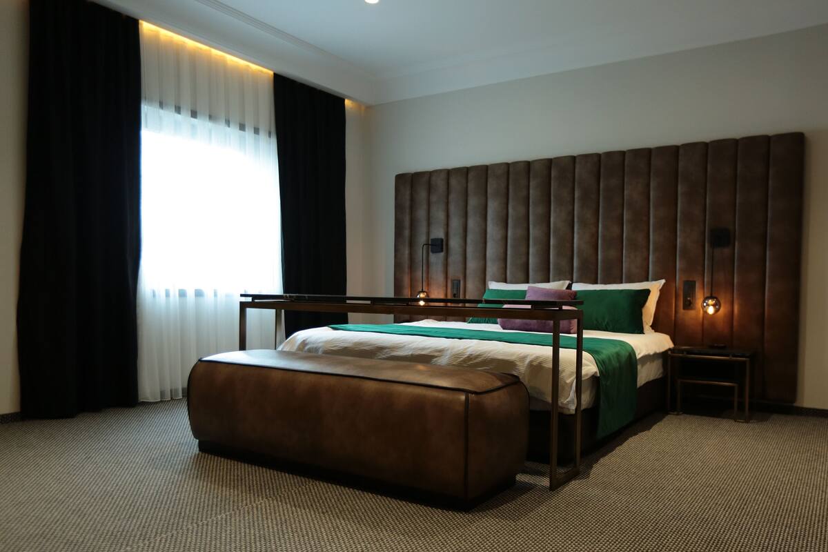 A modern hotel room with a large bed, a wooden headboard, a bench, and a bedside table.