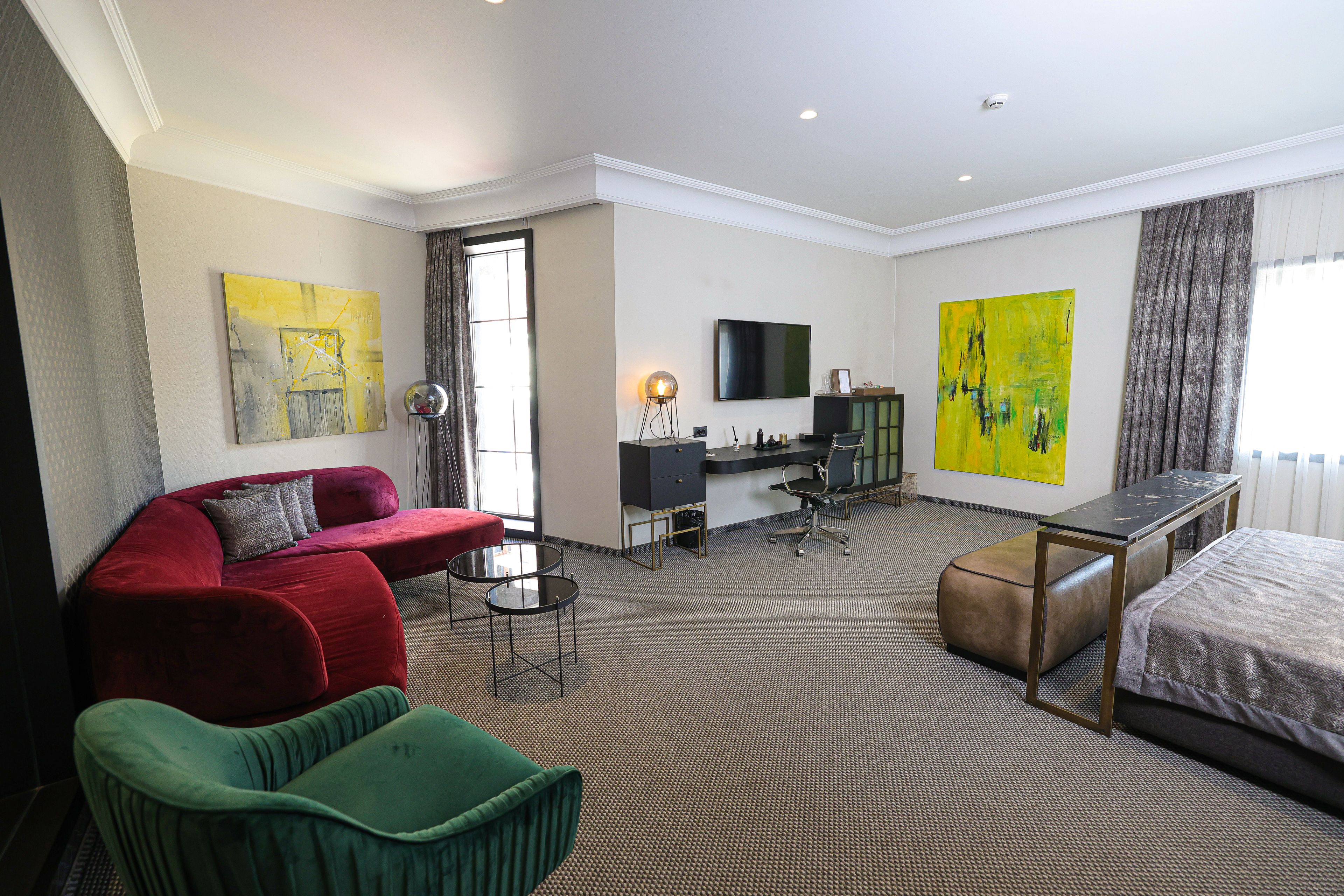 Une chambre d’hôtel avec un canapé rouge, un fauteuil vert, un lit, un bureau avec une chaise, une télévision et deux grands tableaux.