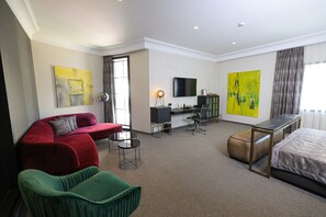A hotel room with a red sofa, green armchair, a bed, a desk with a chair, a TV, and two large paintings. - Hotel Rubis (Ferizaj)