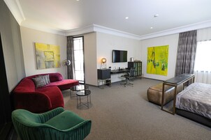 Habitación de hotel con sofá rojo, sillón verde, cama, escritorio con silla, televisor y dos cuadros grandes.