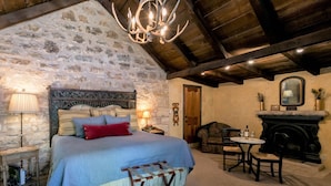 A bedroom with a stone wall, a wooden ceiling, a bed with a headboard, bedside lamps, a small table with chairs, and a fireplace.