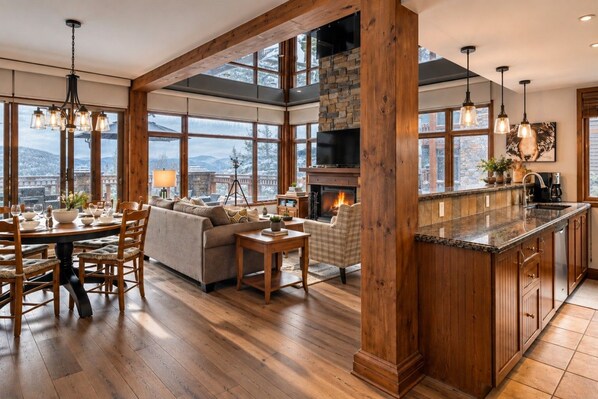 A spacious living area with a fireplace, wooden beams, and large windows offering a view of the outdoors.