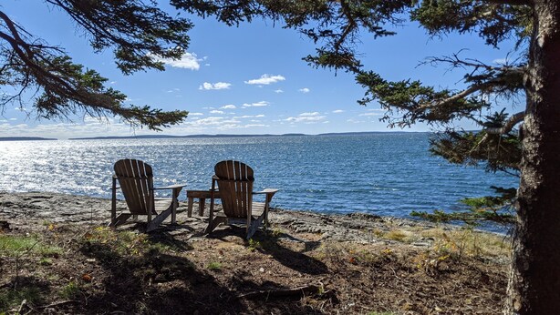 Two Adirondack chairs facing a lake with a rocky shoreline and trees framing the view.