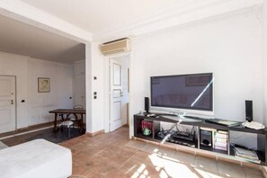 A living room with a flat-screen TV, a coffee table, a sofa, a dining table with chairs, and a door leading to another room.