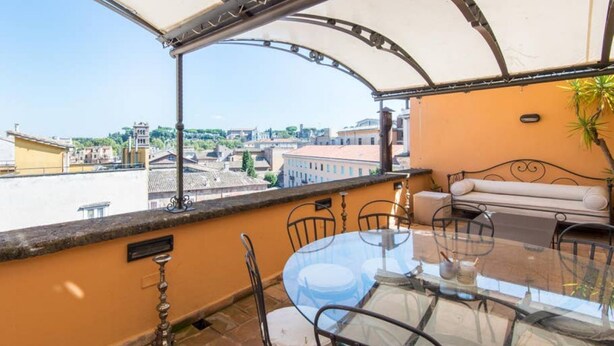 Un balcón con mesa y sillas de vidrio, un sofá y vistas de la ciudad.