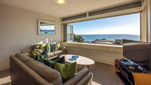 A modern living room with a sofa, coffee table, and a TV on a stand. There is a large window with a view of the sea. - Apartment On The Beach (Waiheke Island)