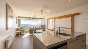 A modern kitchen with a large island, a dining area with a view, and a built-in oven. - Apartment On The Beach (Waiheke Island)