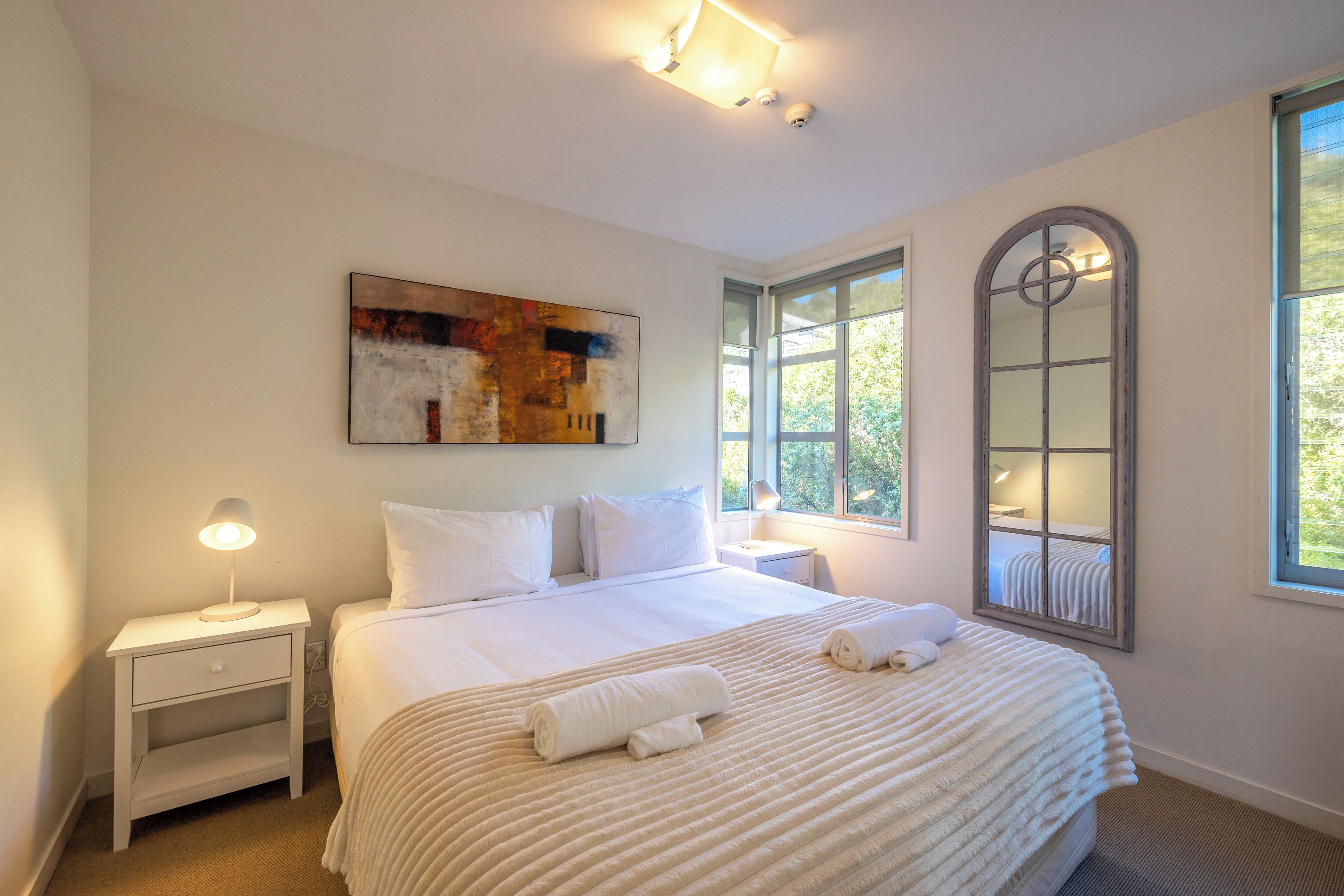 A bedroom with a large bed, a nightstand, a wall-mounted mirror, and a window with natural light.