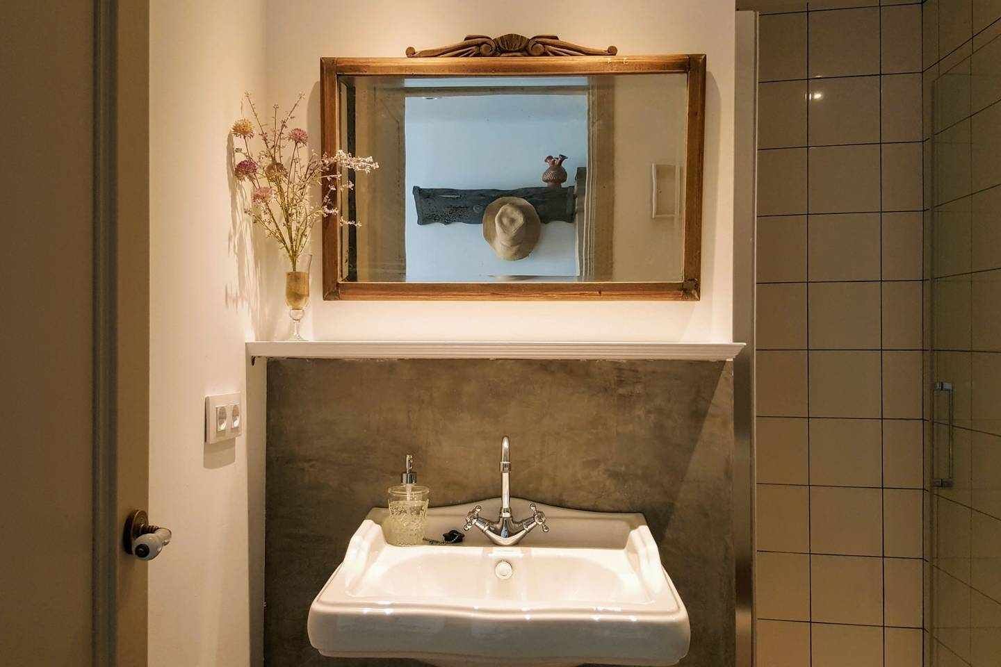Ein Badezimmer mit einem weißen Waschbecken, einem Spiegel darüber und einer Vase mit Blumen auf der linken Seite.