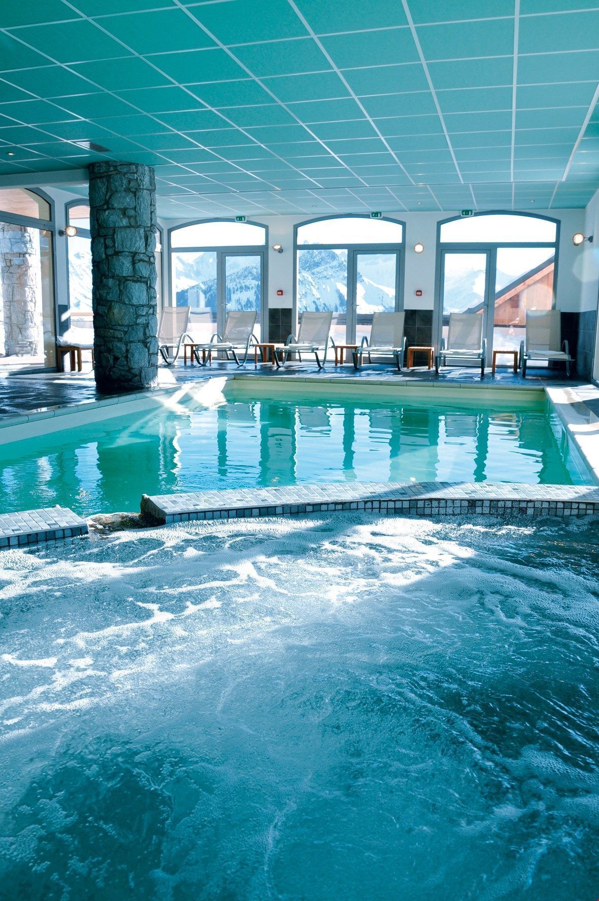 Une piscine intérieure offrant une vue dégagée sur les montagnes grâce à de grandes fenêtres, entourée de chaises longues et d’un pilier en pierre.
