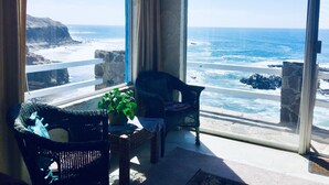 A balcony with wicker furniture, a potted plant, and a view of the ocean and rocky coastline.