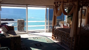 A room with a large sliding glass door leading to a balcony overlooking the ocean. There is a bed with a canopy, a chair, and a small table with a plant.