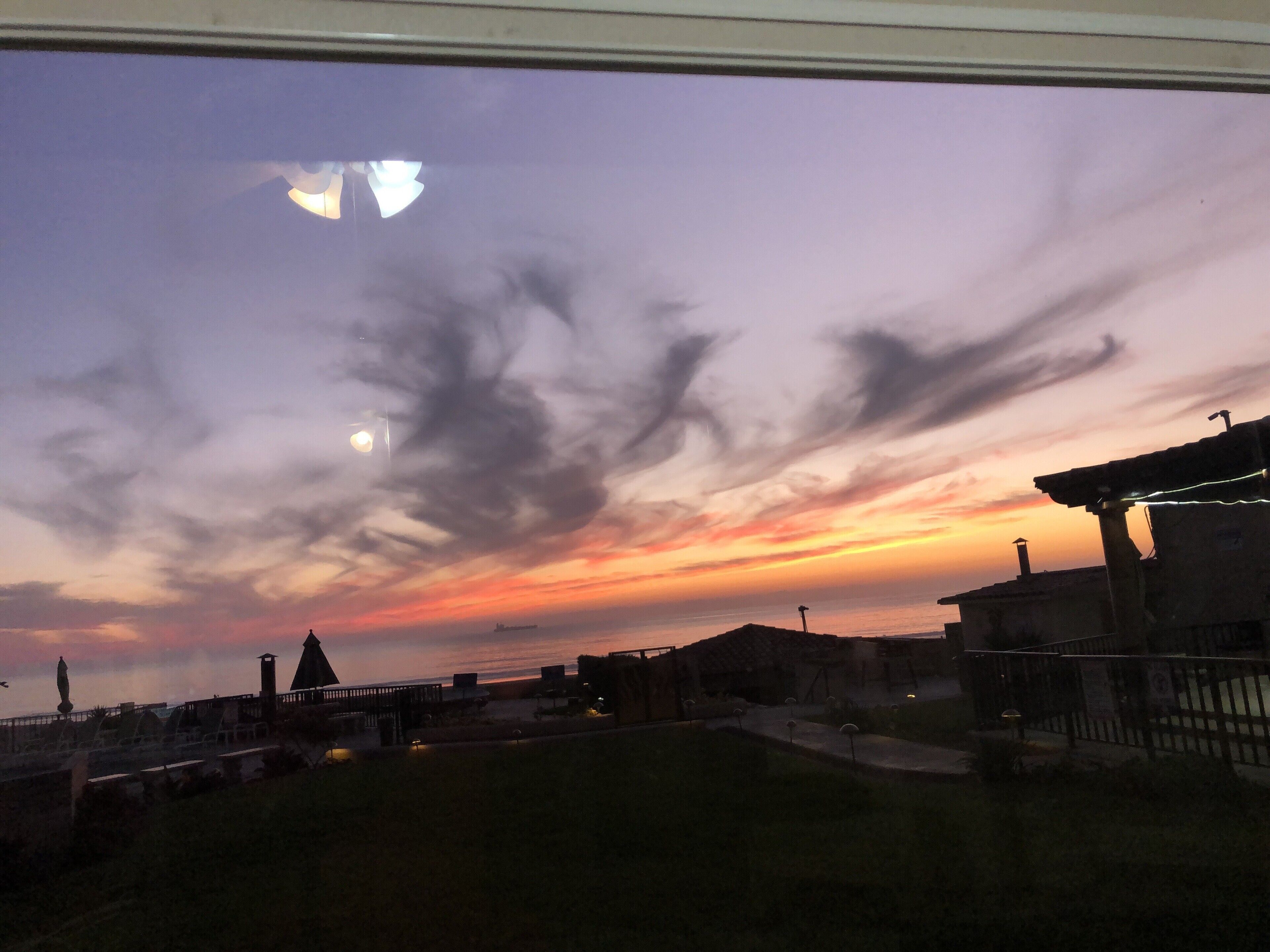 A sunset view from a window overlooking a grassy area with buildings and a distant ship on the horizon.