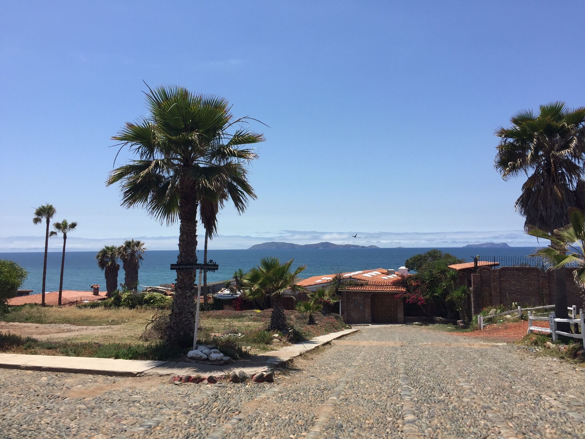A coastal road leading to a building with a tiled roof, surrounded by palm trees and a clear view of the sea and distant mountains.