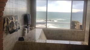 Une salle de bain dotée d’une grande fenêtre donnant sur l’océan, d’une baignoire et d’un mur carrelé où sont disposées des serviettes et des articles de toilette.