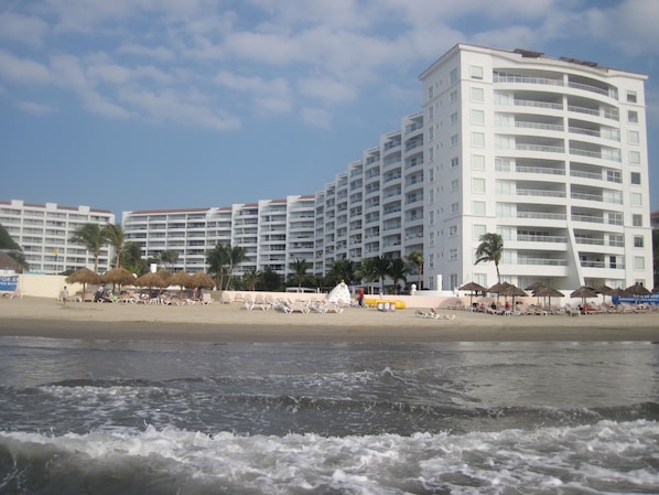 A beachfront hotel with multiple balconies, a sandy beach, and thatched umbrellas.
