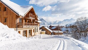 A wooden chalet with a balcony, surrounded by snow-covered mountains and a clear sky.