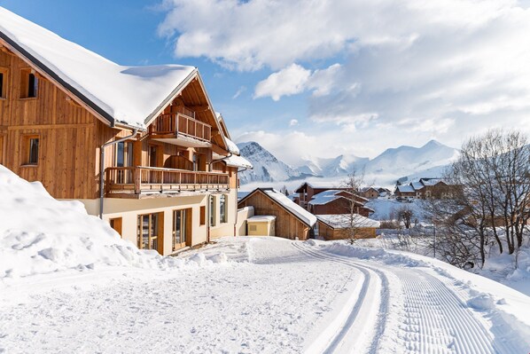A wooden chalet with a balcony, surrounded by snow-covered mountains and a clear sky.