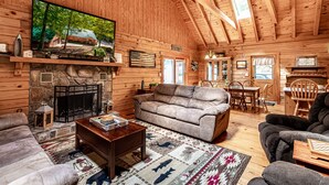 Ein gemütliches Blockhaus-Interieur mit Kamin aus Stein, einem großen Flachbildfernseher an der Wand, einer bequemen Couch und einem Essbereich mit Holzmöbeln.
