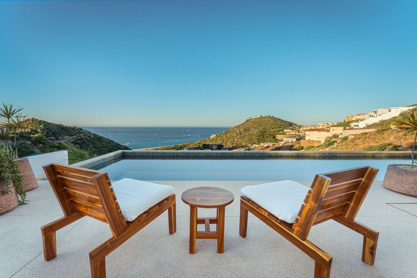 An infinity pool with wooden chairs and a table, overlooking a coastal landscape.