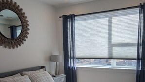 A bedroom with a bed, a ceiling fan, a window with curtains, a nightstand with a lamp, and a mirror with a leaf design.