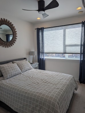 Un dormitorio con cama, ventilador de techo, ventana con cortinas, mesita de noche con lámpara y espejo con diseño de hojas.