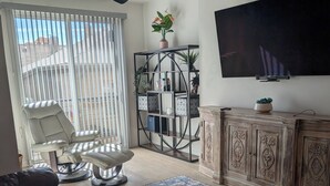 Ein Wohnzimmer mit einer Ledersofa, einem Fernseher, einem Deckenventilator, einem großen Fenster mit Jalousien, einem gemusterten Teppich und einem Holzkasten.