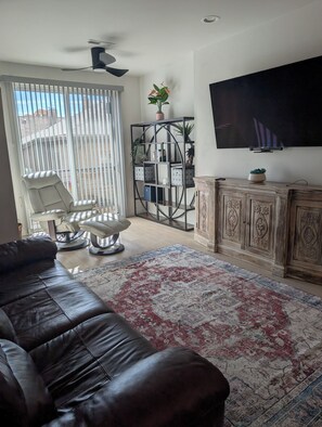 A living room with a leather sofa, a TV, a ceiling fan, a large window with blinds, a patterned rug, and a wooden cabinet.