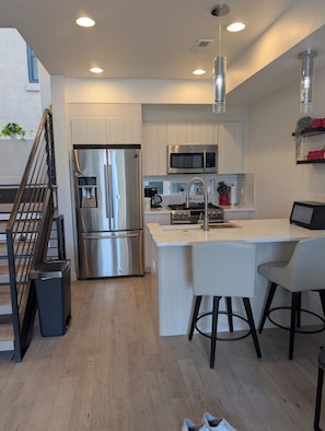 Una cocina moderna con electrodomésticos de acero inoxidable, una isla blanca con taburetes y una escalera con un cubo de basura.