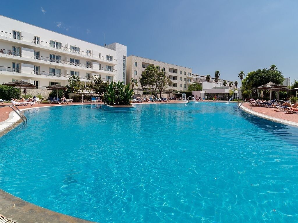 Una amplia piscina al aire libre rodeada de sillones de descanso y sombrillas, con un edificio de hotel de varios pisos al fondo.