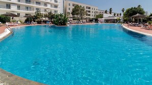 Una amplia piscina al aire libre rodeada de sillones de descanso y sombrillas, con un edificio de hotel de varios pisos al fondo.