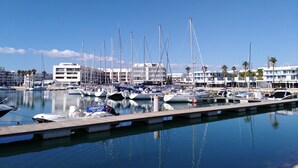 Un puerto deportivo con varias embarcaciones amarradas, un cielo azul despejado y edificios al fondo.