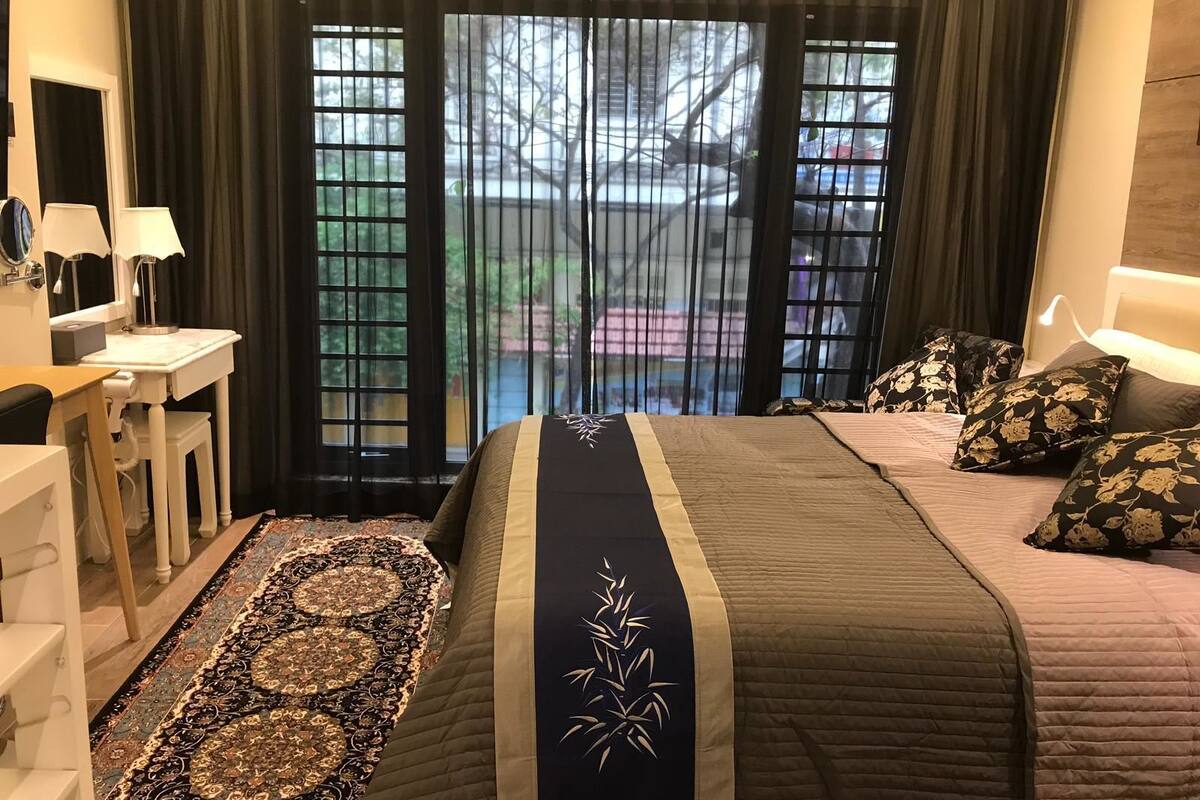 A hotel room with a bed, bedside table, lamp, and a view through a window with a curtain.