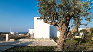 A white building with a stone pathway leading to a white door, surrounded by a garden with various plants and a large tree. - Marza Residence – Bruna - Private villa with pool (Reitani)