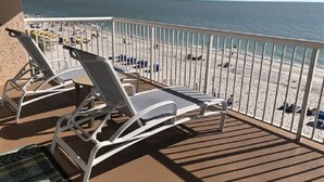 A balcony with white sun loungers and a view of a sandy beach with people and umbrellas.