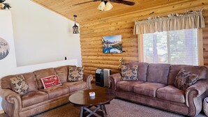 A cozy living room with two brown leather sofas, a wooden ceiling, a ceiling fan, a fireplace, and a window with curtains.