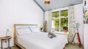 Un dormitorio con cama de madera, una lámpara de pie, un ventilador y una ventana con vistas a la vegetación.