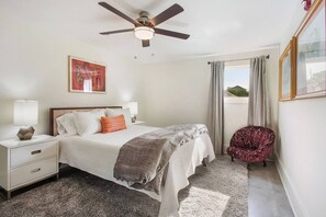 Une chambre à coucher comprenant un lit, des tables de chevet, une chaise, un ventilateur de plafond et une fenêtre avec des rideaux.